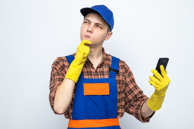 Young cleaning guy wearing uniform and cap with gloves holding phone isolated on white wall