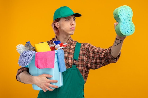 Young cleaning man in plaid shirt jumpsuit and cap holding sponge and bucket with cleaning tools looking aside with serious face ready for cleaning standing on orange