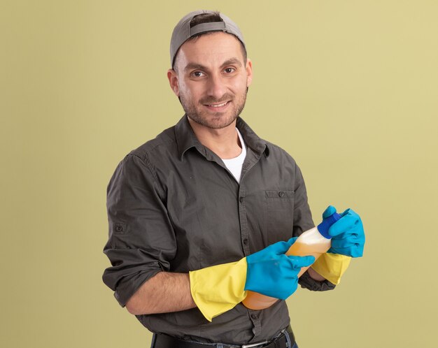 Young cleaning man wearing casual clothes and cap in rubber gloves holding bottle with cleaning supplies looking  with smile on face standing over green wall