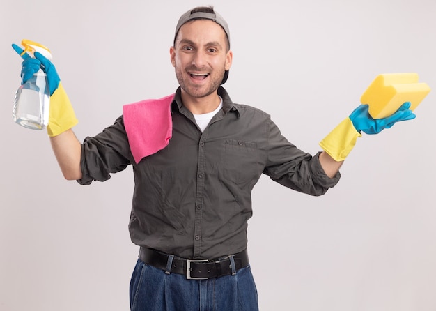 Young cleaning man wearing casual clothes and cap in rubber gloves holding cleaning spray and sponge with rag on his shoulder looking  happy and excited standing over orange wall