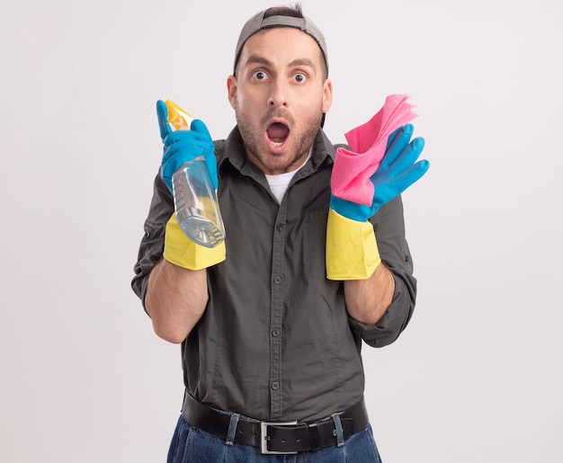 Free photo young cleaning man wearing casual clothes and cap in rubber gloves holding spray bottle and rag looking  amazed and surprised standing over white wall