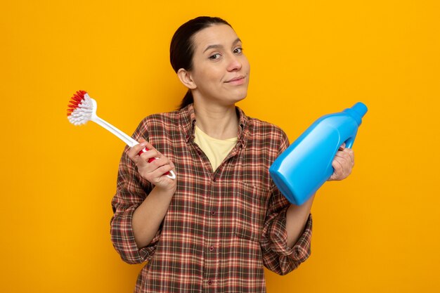 Young cleaning woman in casual clothes holding cleaning brush and bottle of cleaning supplies  with skeptic smile on face standing over orange wall