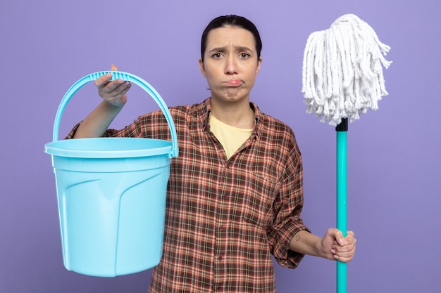 Free Photo young cleaning woman in casual clothes holding mop and bucket  confused and very disappointed making wry mouth standing over purple wall