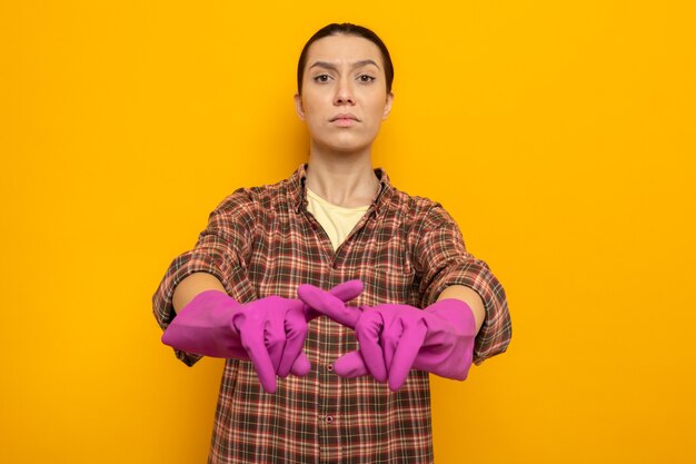 Young cleaning woman in casual clothes in rubber gloves looking with serious face making defense gesture crossing index finger