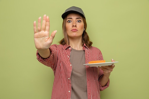 Young cleaning woman in plaid shirt and cap holding plate and sponge looking at camera with serious face making stop gesture with hand standing over green background