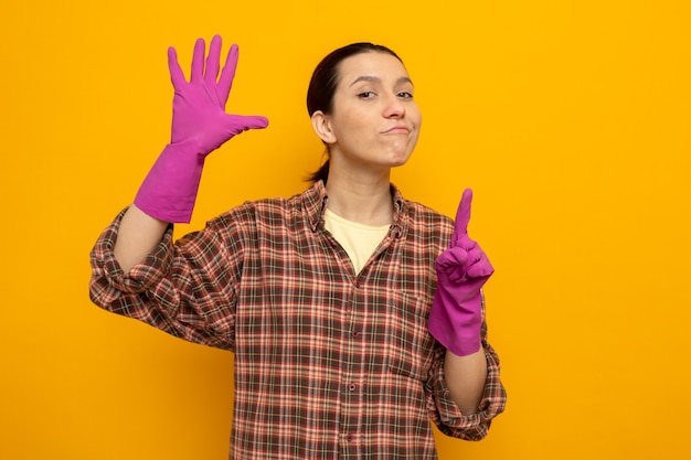 Young cleaning woman in plaid shirt in rubber gloves with confident smile on face showing number six with fingers standing on orange