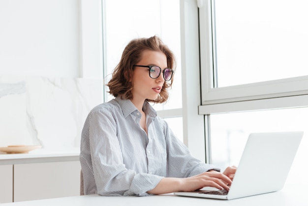 young concentrated woman in striped shirt using laptop while siting at table in light apartment