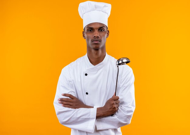 Young confident afro-american cook in chef uniform crosses arms and holds ladle isolated on orange background with copy space