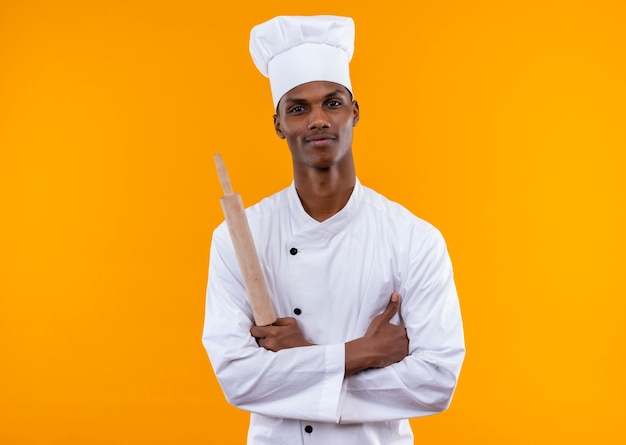 Young confident afro-american cook in chef uniform crosses arms and holds rolling pin isolated on orange background with copy space