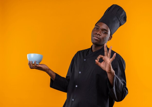 Young confident afro-american cook in chef uniform holds bowl and gestures ok hand sign on orange  with copy space