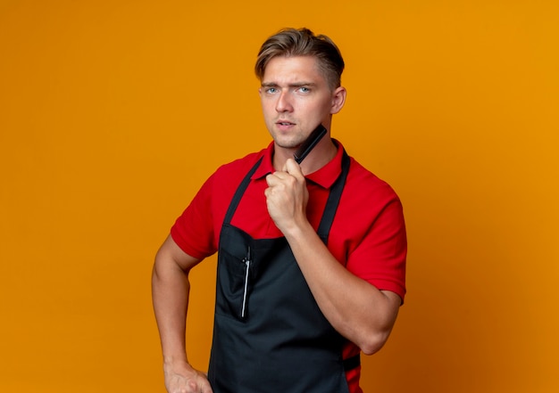 Young confident blonde male barber in uniform holds and puts comb on chin isolated on orange space with copy space