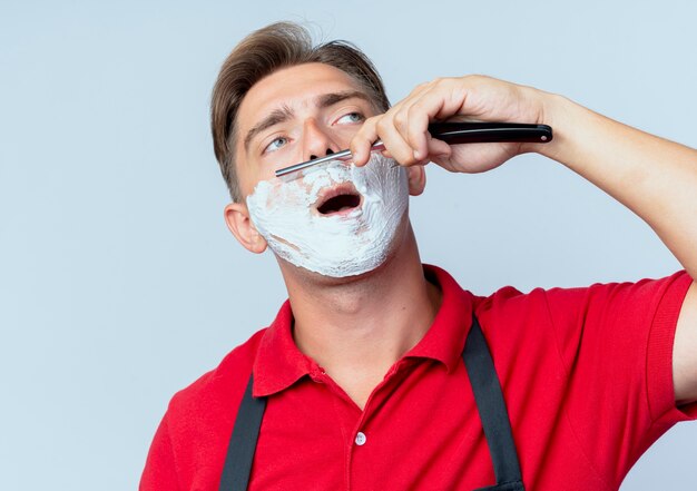 Young confident blonde male barber in uniform smeared face with shaving foam shaving straight razor looking at side isolated on white space with copy space