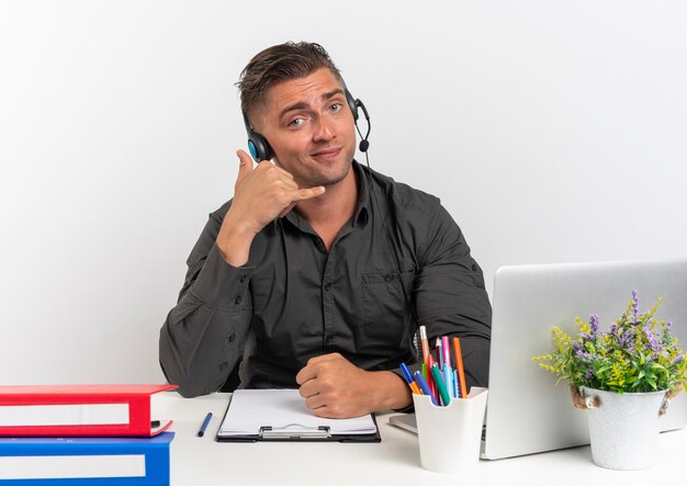 Young confident blonde office worker man on headphones sits at desk with office tools using laptop gestures phone isolated on white background with copy space