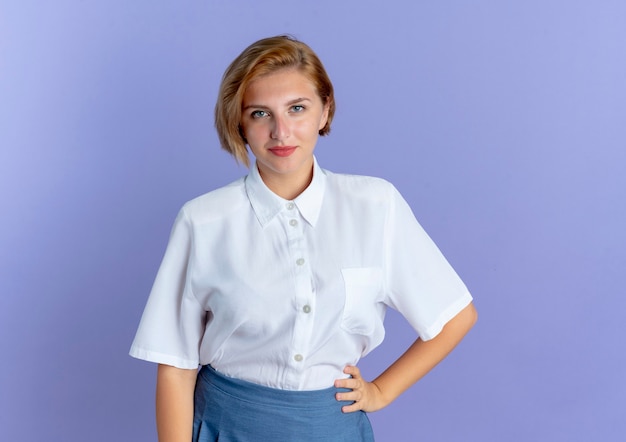 Young confident blonde russian girl puts hand on waist looking at camera isolated on purple background with copy space