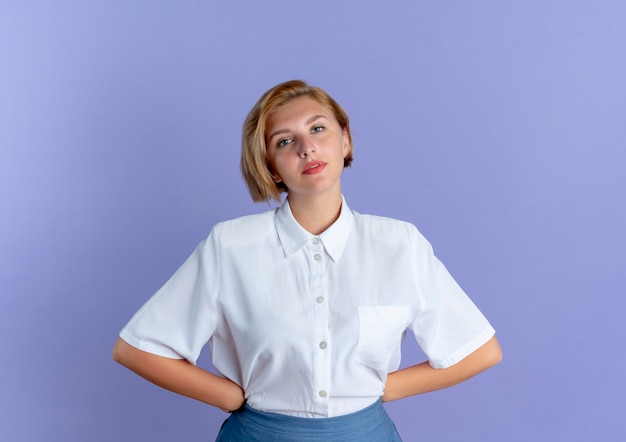 Young confident blonde russian girl puts hands on waist looking at camera isolated on purple background with copy space