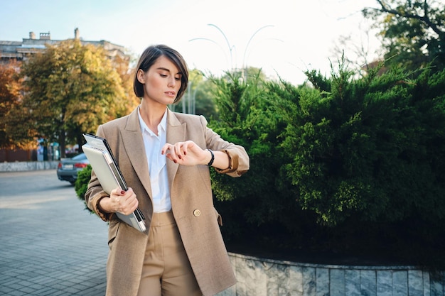 Young confident businesswoman in beige suit standing with laptop watching time on wristwatch outdoor