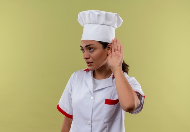 Young confident caucasian cook girl in chef uniform gestures can't hear sign isolated on green wall with copy space