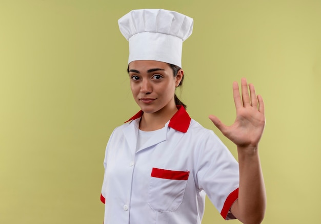 Young confident caucasian cook girl in chef uniform gestures stop hand sign isolated on green wall with copy space