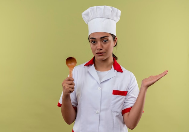 Young confident caucasian cook girl in chef uniform holds wooden spoon and keeps hand open isolated on green wall with copy space