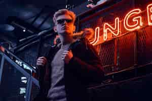 Free photo young confident guy wearing coat with fur hood and sunglasses standing on the staircase to the underground nightclub, a backlit signboard in the background