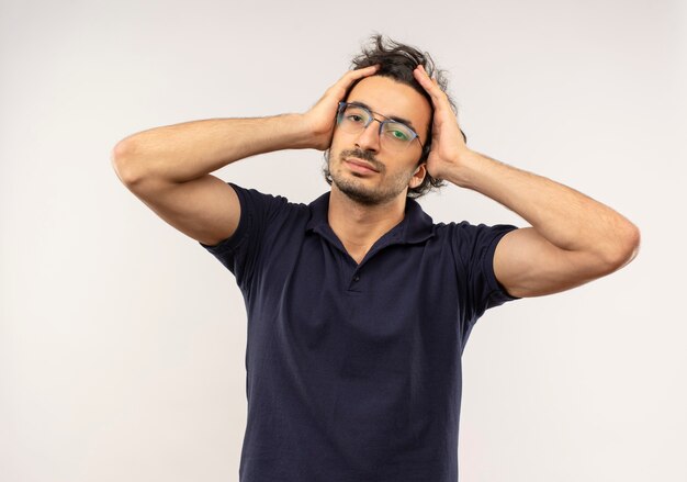 Young confident man in black shirt with optical glasses holds head with hands and looks isolated on white wall