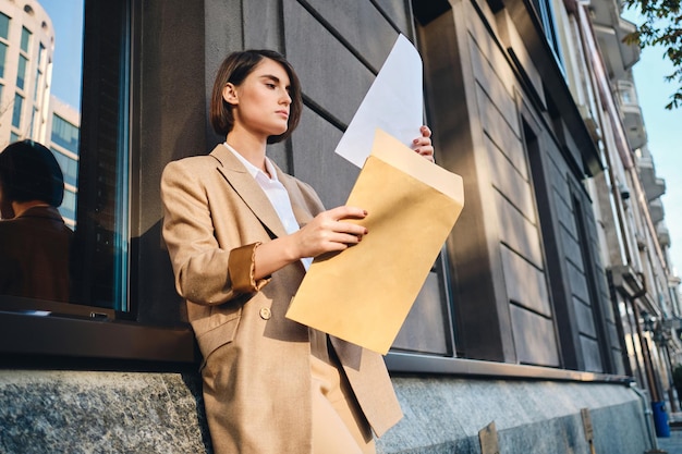 Free photo young confident stylish businesswoman in suit thoughtfully working with papers on city street