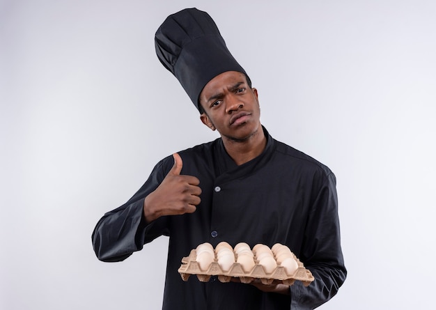 Young confused afro-american cook in chef uniform holds batch of eggs and thumbs up isolated on white wall 