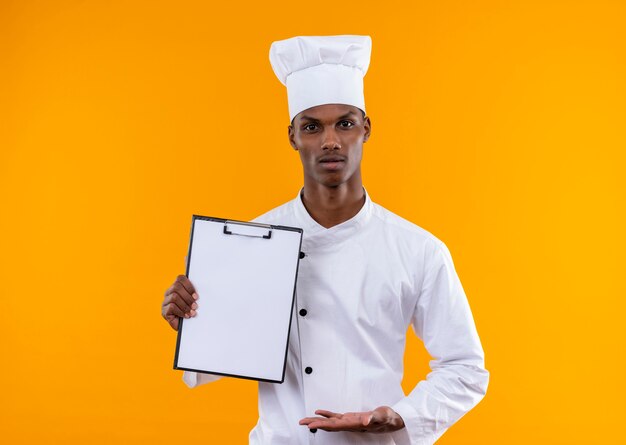 Young confused afro-american cook in chef uniform holds clipboard and holds hand open isolated on orange wall