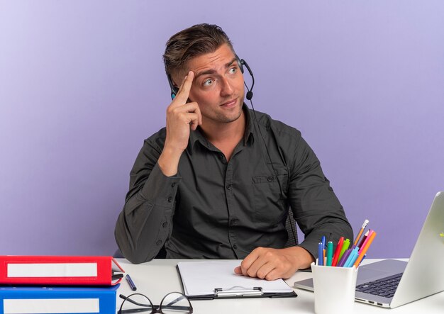 Young confused blonde office worker man on headphones sits at desk with office tools using laptop  puts hand on head looking at side isolated on violet space with copy space