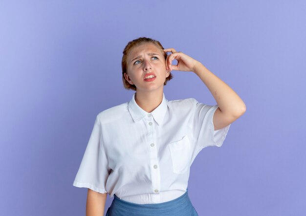 Young confused blonde russian girl puts hand on head isolated on purple background with copy space