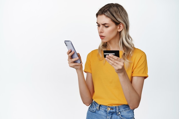 Free Photo young confused woman holding credit card and looking at smartphone screen with puzzled face, concept of problem with money transfer or payment in app on white.