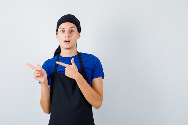 Young cook in t-shirt, apron pointing left and looking shocked , front view.