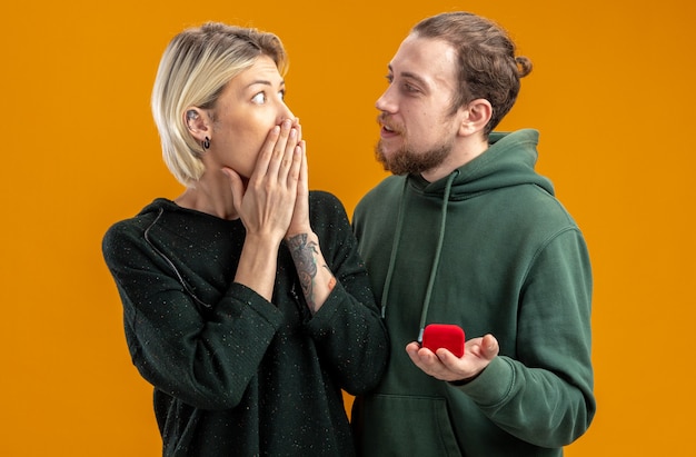 Free Photo young couple in casual clothes happy man making proposal with engagement ring in red box to his amazed and surprised girlfriend valentines day concept standing over orange wall