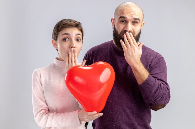 young couple in casual clothes man and woman holding heart shaped balloon looking at camera amazed and surprised covering mouth with hands celebrating valentines day standing over white background