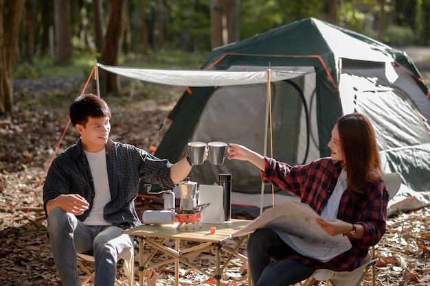 Free photo young couple clinking coffee mugs together in the morning in front of a camping tent in the morning at nature park