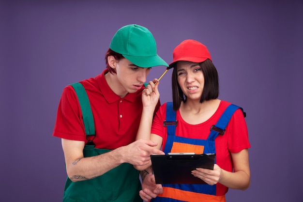 Young couple in construction worker uniform and cap confused girl holding pencil and clipboard  touching head with pencil concentrated guy looking and pointing at clipboard isolated