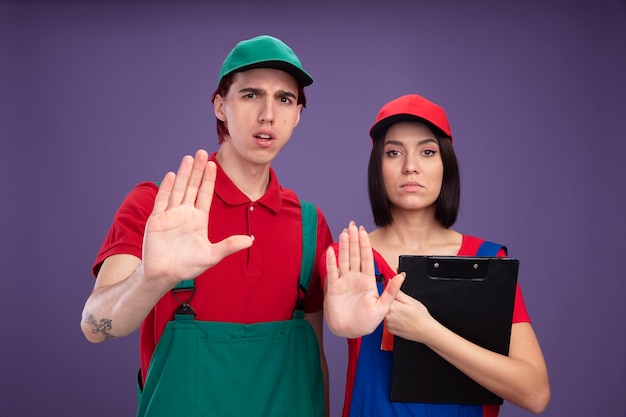 Young couple in construction worker uniform and cap frowning guy serious girl holding pencil and clipboard both looking at camera doing stop gesture isolated on purple wall