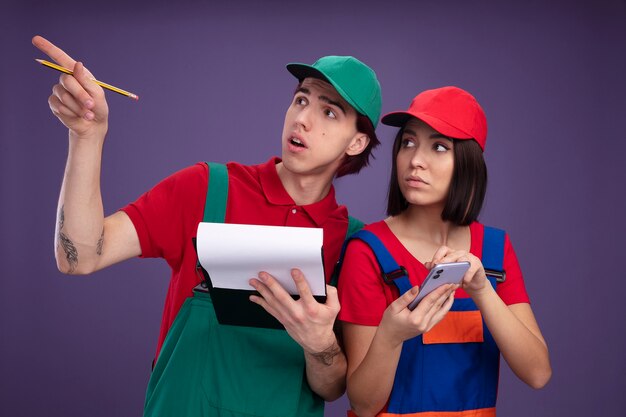 Young couple in construction worker uniform and cap impressed guy holding pencil and clipboard pointing up serious girl holding mobile phone both looking up isolated on purple wall