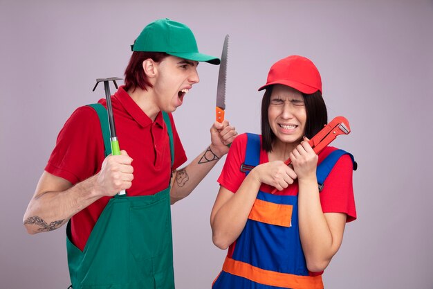 Young couple in construction worker uniform and cap scared girl holding pipe wrench with closed eyes angry guy holding hoe-rake and hand saw looking at girl shouting 