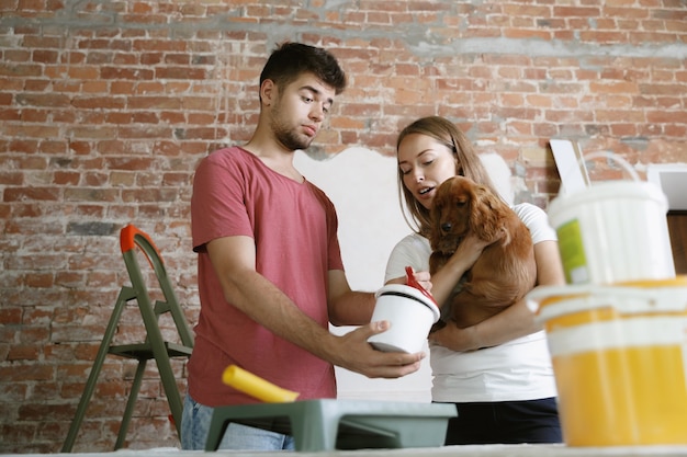 Free Photo young couple doing apartment repair together themselves. married man and woman doing home makeover or renovation. concept of relations, family, pet, love. choosing the paint color, holding the dog.