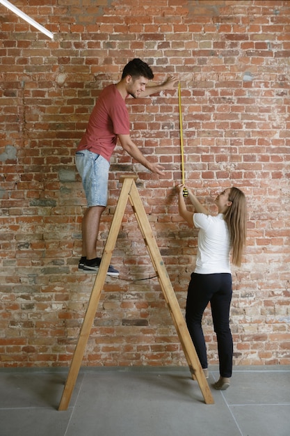 Free photo young couple doing apartment repair together themselves. married man and woman doing home makeover or renovation. concept of relations, family, pet, love. measuring standing on ladder with the metr.