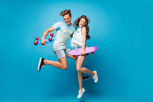 Young couple of handsome guy and pretty girl with long curly hair is jumping on blue background in studio. They wear T-shirts, jeans short, hold skateboards in hands.