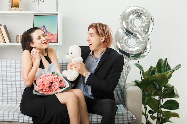 Young couple on happy women day holding teddy bear with bouquet sitting on sofa in living room