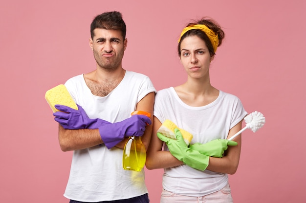 Young couple having frowning faces keeping their hands crossed holding brush, detergent and mops wearing protective gloves and white casual T-shirts having bad mood and unwillingness to clean house