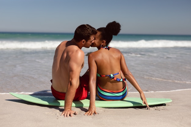 Young couple kissing each other while sitting on surfboard at beach in the sunshine