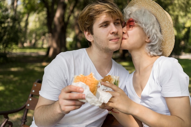 Free photo young couple kissing while eating burgers in the park