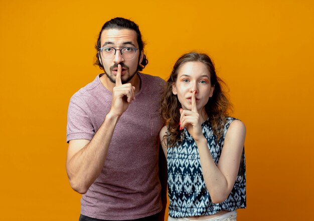 Young couple looking at camera making silence gesture with fingers on lips standing over orange background