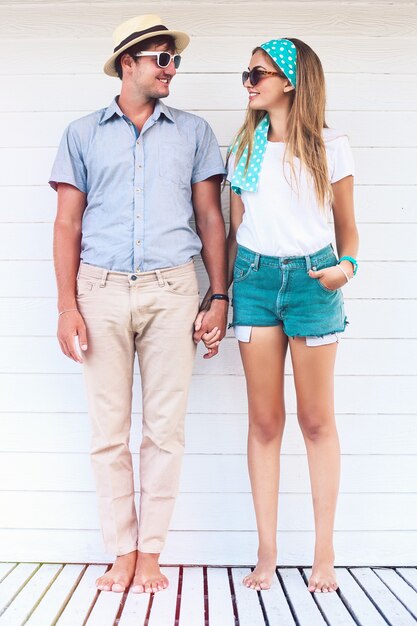 Young couple in love posing near white beach cafe in summer retro bright outfits, holding hands