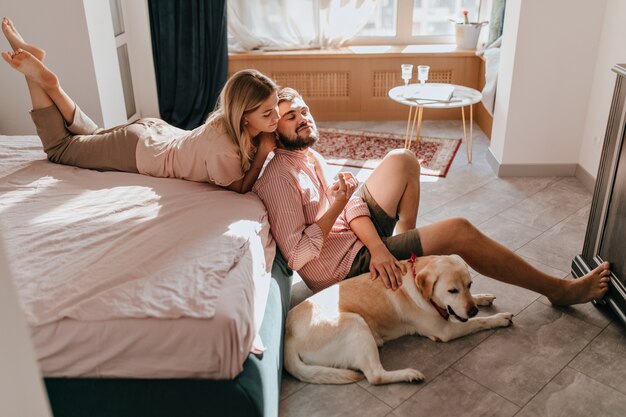 Young couple of lovers resting in bedroom. Girl in comfortable clothes lies on bed and looks at her boyfriend while he is stroking Labrador.