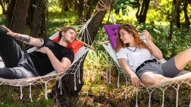 Free photo young couple lying on hammocks, looking at each other and smiling. greenery around. glamping